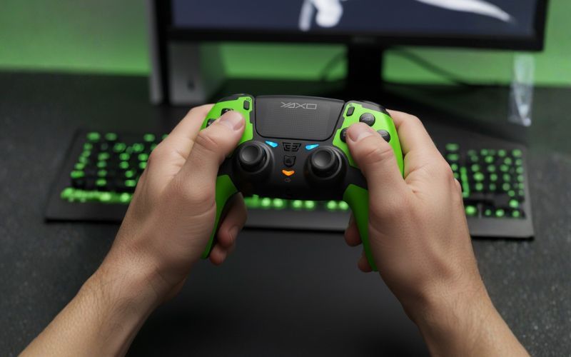 Osoba trzyma zielono-czarny kontroler YAXO Hornet Fury Elite na tle monitora i podświetlanej klawiatury