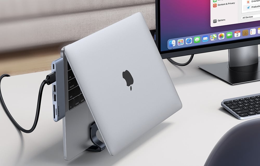 MacBook w podstawce podłączony kablem do stacji dokującej na białym biurku z monitorem i klawiaturą