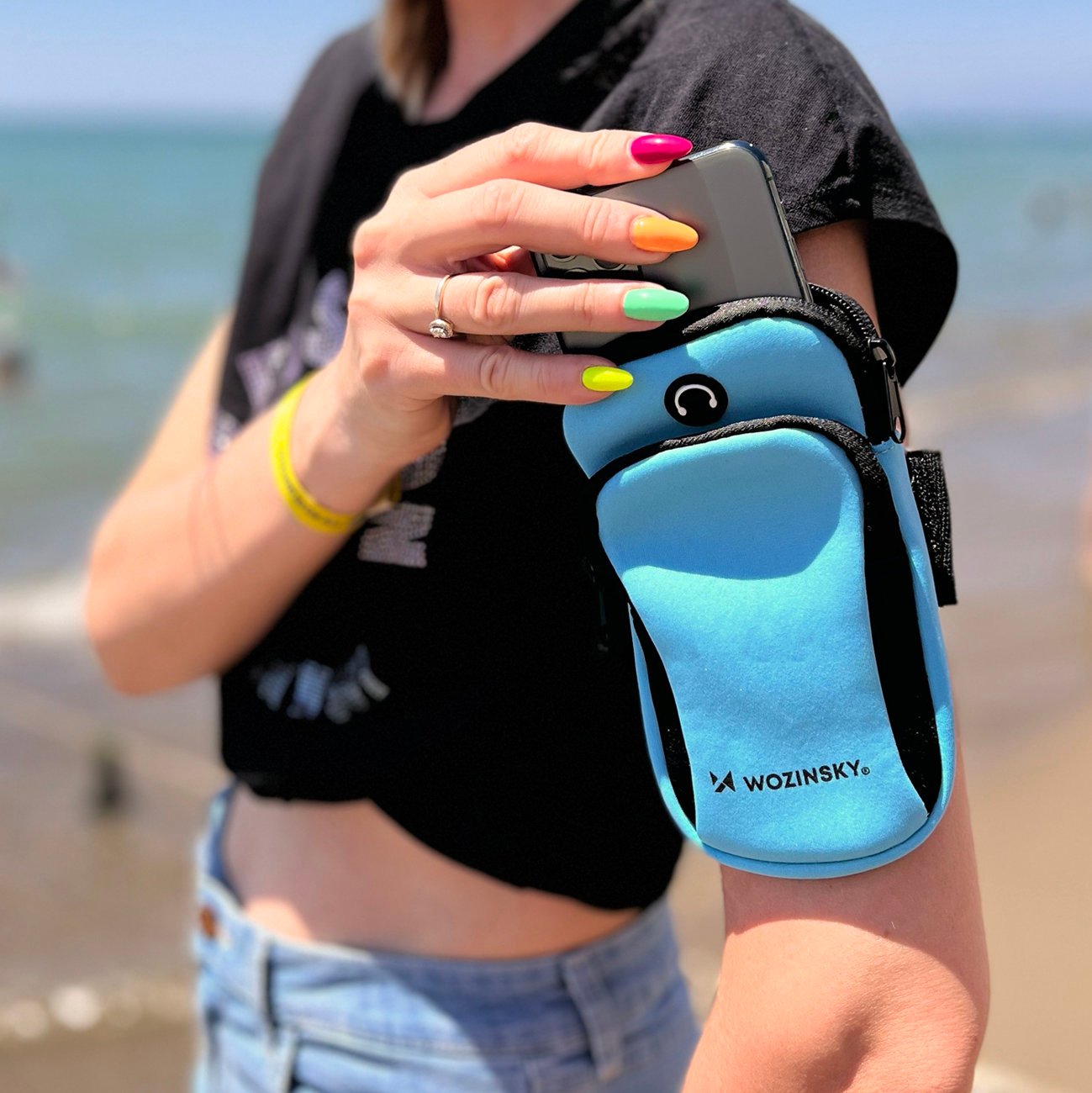 Niebieska opaska do biegania na telefon Wozinsky WABBL1 pokazywana przez modelkę na plaży Tak prezentuje się niebieska opaska do biegania na telefon Wozinsky WABBL1 założona przez modelkę na plaży