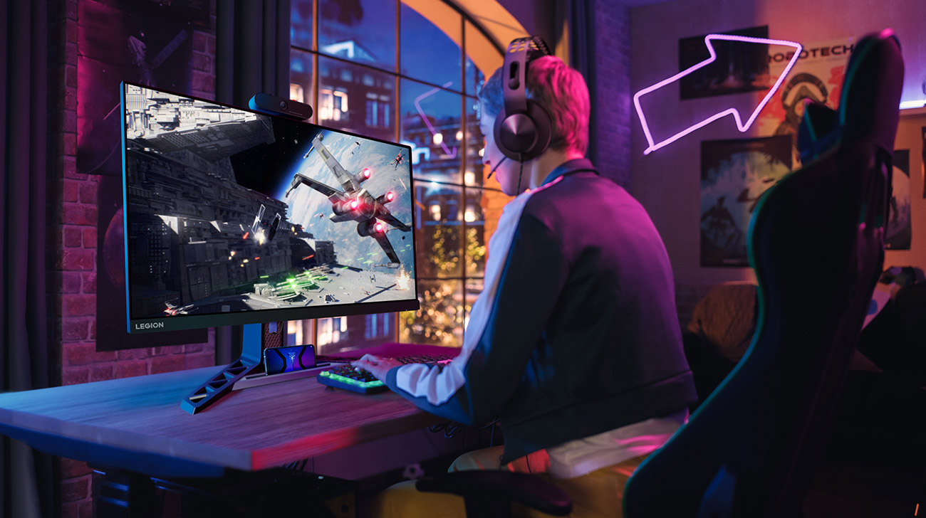 Monitor Lenovo R27q-30 na biurku przed którym siedzi młoda osoba ze słuchawkami na głowie i gra w grę komputerową