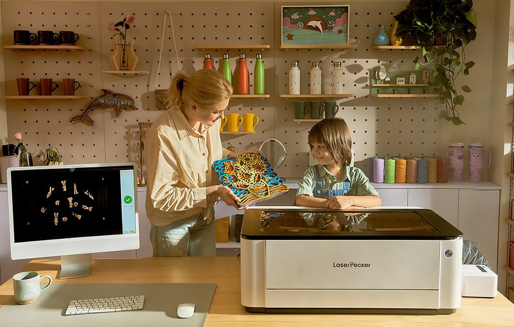 Kobieta pokazuje dziecku kolorową wycinankę przy maszynie LaserPecker na tle półek z kubkami i sznurkami