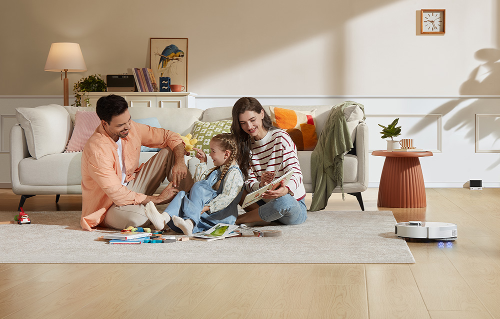 Rodzina z dzieckiem bawi się na dywanie w salonie. Po prawej stronie robot czyści podłogę.