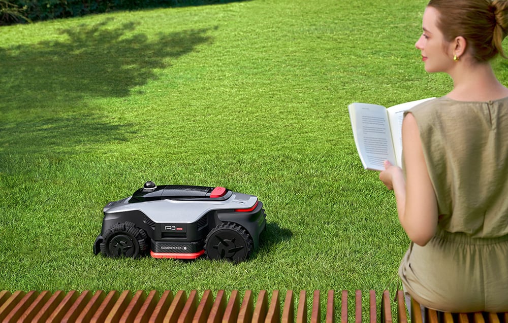 Kobieta czyta książkę na ławce obok pracującego robota koszącego