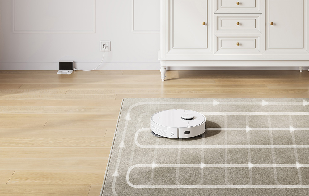 Robot sprzątający odkurza dywan. W oddali widoczna jest stacja dokująca podłączona do gniazdka.