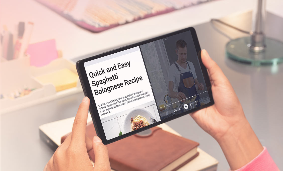 Tablet Lenovo Tab One trzymany w dłoniach, na ekranie przepis kulinarny, w tle biurko z akcesoriami biurowymi