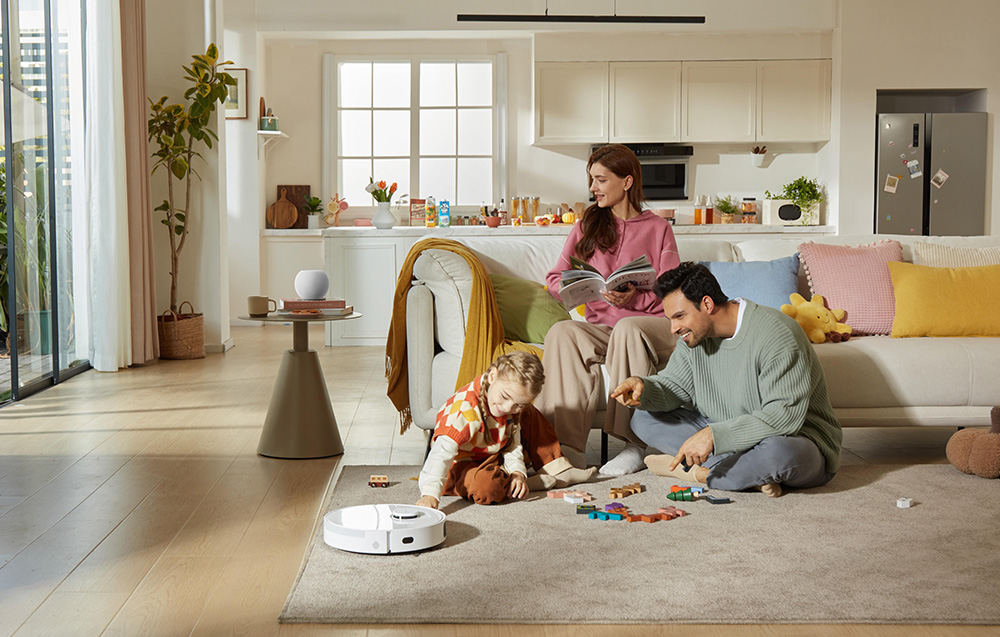 Rodzina z dzieckiem bawi się w jasnym salonie. Robot sprzątający odkurza dywan, na stoliku stoi asystent głosowy.