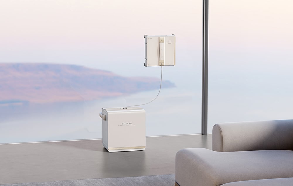 Robot do mycia okien Dreame C1 Station przymocowany do dużej szyby z widokiem na krajobraz, połączony kablem z stacją stojącą na podłodze