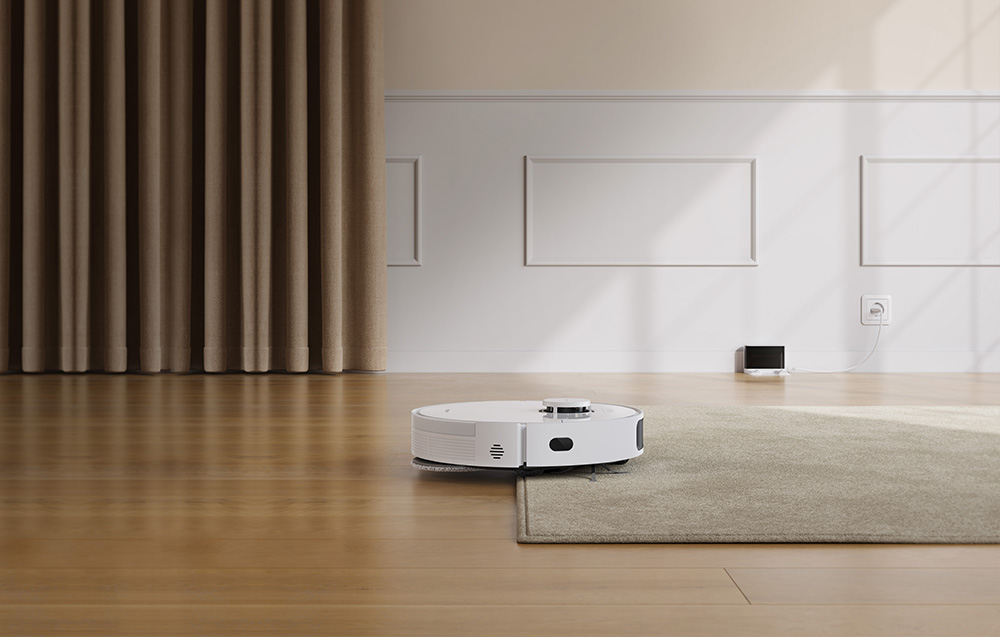 Robot sprzątający odkurza dywan. W oddali stacja dokująca.