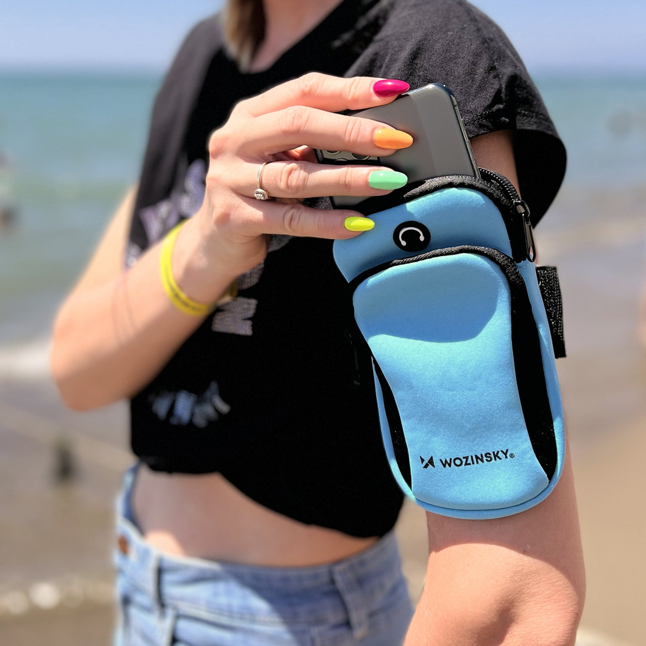 Niebieska opaska do biegania na telefon Wozinsky WABBL1 pokazywana przez modelkę na plaży Tak prezentuje się niebieska opaska do biegania na telefon Wozinsky WABBL1 założona przez modelkę na plaży